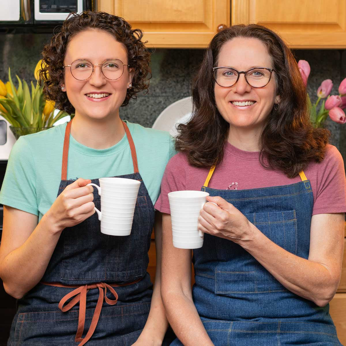 two ladies smiling in a kitchen while holding coffee cups.